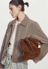 Light brown, textured jacket with a collared design and large buttons. Brown suede bag with silver detailing, held in one hand.