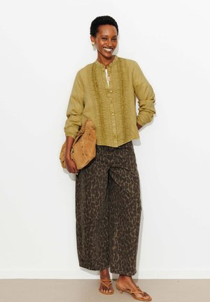 Femme souriante portant un chemisier olive texturé, un pantalon large à imprimé léopard, tenant un sac en daim beige, et des sandales à lanières, sur fond blanc.