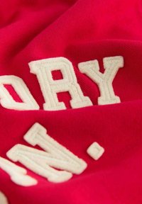 Červená látka s vyvýšenými, vyšívanými bílými písmeny, které hlásají "DRY" a "N." Měkká textura, mírně pomačkaný vzhled díky skládání.