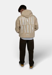 Felpa con cappuccio beige con grande testo stampato bianco "CRYSTAL PARIS" sul retro, abbinata a pantaloni scuri e sneakers.
