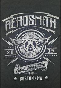 Černé tričko s vybledlým šedým potiskem se slovy "AEROSMITH," křídly, hvězdami a "Aero Force One" kolem kruhového emblému.