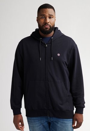 Schwarze Zip-Hoodie mit einer Fronttasche und Kordelzug, versehen mit einem kleinen Logo auf der Brust. Kombiniert mit blauen Jeans.
