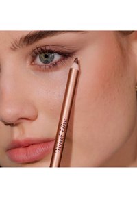 Matita per labbra color rosé dorato tenuta vicino alla bocca di un modello; presenta una punta appuntita e la scritta "Velvet Love" incisa sul fusto.