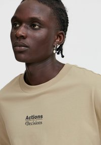 Beige cotton t-shirt with short sleeves and crew neck, featuring the printed text "Actions and Decisions" in dark brown.