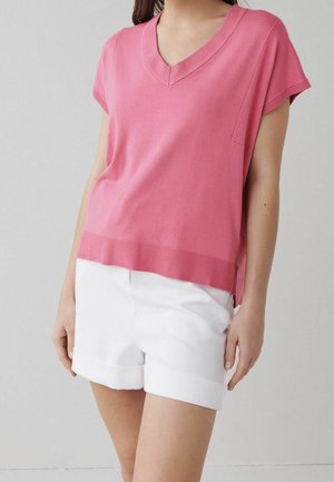 Frau trägt einen lockeren pinkfarbenen V-Ausschnitt mit kurzen Ärmeln und weiße gekrempelte Shorts, steht vor einem schlichten hellen Hintergrund.