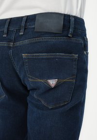 Krupni plan tamnoplavih traperica s naborom na stražnjem džepu, s narančastim šavovima i crveno-bijelim logotipom u obliku trokuta Guess.