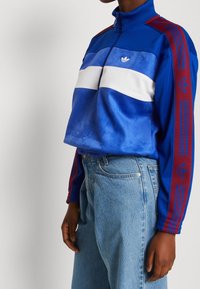 Person som bär en blå Adidas-träningsjacka med röda ränder och Trefoil-logotyper på ärmarna, i kombination med högmidjade ljusblå jeans.