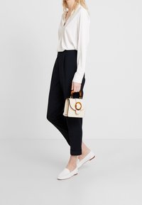 Chemise blanche à manches longues, pantalon bleu marine fuselé, chaussures blanches sans lacets, tenant un petit sac à main crème avec une poignée circulaire marron.
