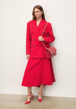 Piros, dupla soros zakó nagy fekete gombokkal, hozzáillő szoknyával párosítva. Piros pántos magassarkúk. Kiegészítőként piros kígyóbőr kézitáska.