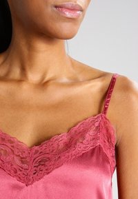 Mujer vestida con un top de satén rosa con encaje, mostrando el escote, los hombros y parte de la cara contra un fondo liso.