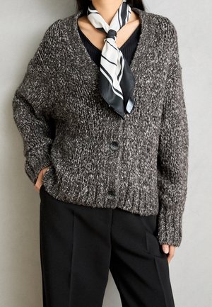 Persoon die een dikke grijze gebreide cardigan met zwarte knopen draagt, een zwart topje, zwarte broek en een zwart-witte gestreepte halsdoek.