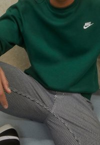 Grön sweatshirt med rund halsringning och vitt Nike-logga; matchat med svart-vit randiga byxor. Synlig hand som vilar på låret.