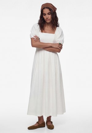 Kobieta w długiej białej sukience z bufiastymi rękawami, brązową chustą na głowie i pasującymi brązowymi butami Mary Jane, stojąca z założonymi ramionami.