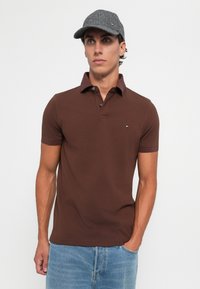 Ung mann med grå caps, brun kortermet polo skjorte og lyseblå jeans, står med den ene hånden i lomma mot hvit bakgrunn.