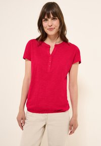 Haut rouge à manches courtes avec un col rond et une patte à trois boutons. Texture douce avec une coupe décontractée et un ourlet subtil.