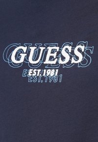 Granatowa bawełniana koszulka z białym i jasnoniebieskim haftowanym logo "GUESS" oraz tekstem "EST. 1981", o miękkiej fakturze.