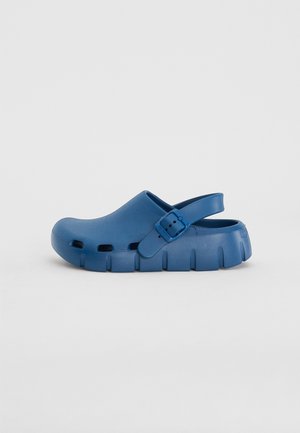 Blåt slip-on træsko med justerbar bagrem, ventilerede sider og tyk struktureret sål, vist i profil mod en neutral baggrund.