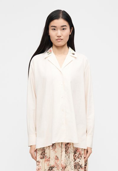 Jovem com cabelos escuros e longos, vestindo uma camisa creme de botões larga com bordado floral na gola e uma saia plissada com padrão floral.