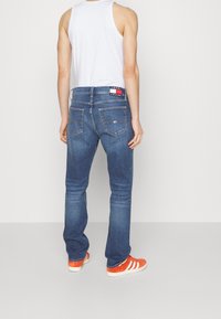 Vaqueros de mezclilla azul con un corte relajado, costuras visibles, bolsillos traseros y una etiqueta de marca en rojo, blanco y azul en la cintura. Llevados con zapatillas naranjas.
