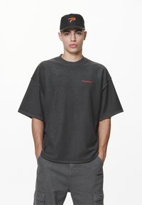 Ungdommelig mann med svart caps med oransje logo, mørk oversized kortermet skjorte og matchende cargo-bukser, stående med hendene i lommene.