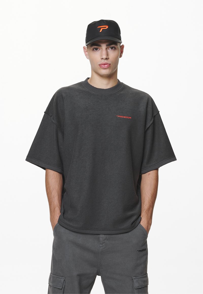 Ungdommelig mann med svart caps med oransje logo, mørk oversized kortermet skjorte og matchende cargo-bukser, stående med hendene i lommene.