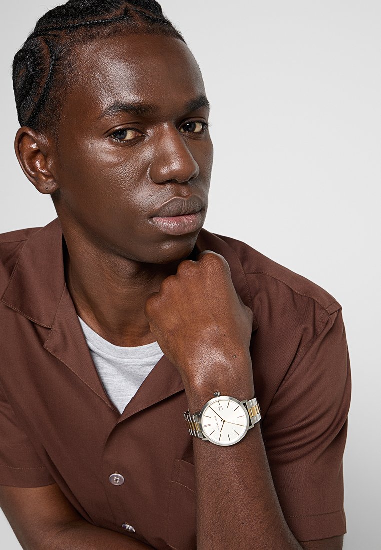 Montre-bracelet en argent et or avec un cadran blanc, dotée de marqueurs horaires fins et d'un affichage de date subtil, portée sur un poignet à la peau foncée.