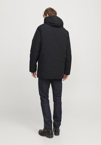 Sort polstret jakke med hætte, der har en glat overflade og en lige pasform. Båret over mørke denim jeans og sorte støvler.