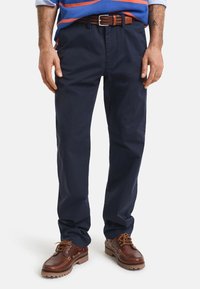 Pantalones de algodón azul marino, diseño de pierna recta, con bolsillos delanteros. Combinados con zapatos marrones con cordones y suela robusta. Cinturón trenzado marrón como accesorio.