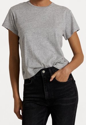 Personne portant un t-shirt gris chiné à manches courtes légèrement rentrée dans un jean noir taille haute, une main dans la poche.