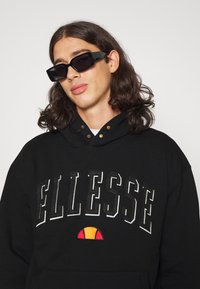 Zwarte hoodie met het geborduurde "ELLESSE" logo, met een oranje en rode accent eronder. Vergezeld van vierkante, zwarte zonnebril.