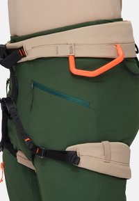 Groene klimspijkerbroek met een beige tailleband, voorzien van een ritszak, verstelbare banden en een oranje karabijnhaakbevestiging.