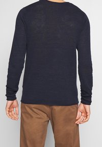 Osoba v modrém svetr s dlouhým rukávem a hnědých kalhotech, zobrazená zezadu na jednolitém pozadí.