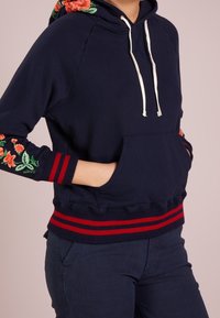 Marinblå hoodie med blommönster broderat på ärmarna, vita snörningar och röda randiga manschetter och midja. Framtida kängurufickor.