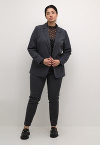 Uhlově šedý oblek se střiženým sakem a slim-fit kalhotami. Pod ním je průhledný černý top s puntíky. Černé mokasíny se zlatými akcenty.