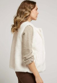 Gilet en fausse fourrure blanche avec un col montant et des poches latérales, associé à un pull beige à manches retroussées. Texture lisse.