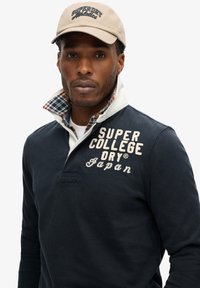 Marineblauwe longsleeve polo met geborduurde tekst "SUPER DRY", witte kraag en geruite voering. Beige pet met het logo "Superdry Athletics".
