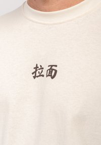 Prim-plan cu o cămașă bej deschis cu caractere chinezești întunecate tipărite în centrul zonei pieptului.