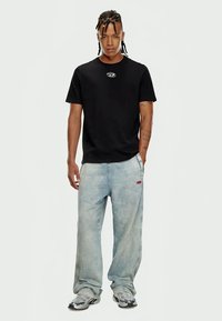 Man with braided hair wearing black t-shirt, light blue baggy jeans, and silver sneakers, standing with one hand in pocket.