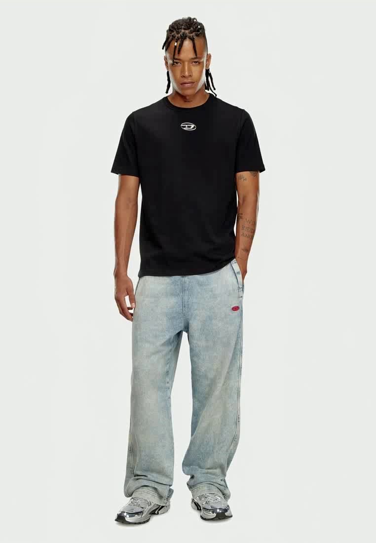 Homme avec des cheveux tressés portant un t-shirt noir, un jean baggy bleu clair et des baskets argentées, se tenant avec une main dans la poche.