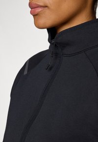 Veste zippée noire à col montant, avec une texture lisse, des coutures visibles et des accents gris subtils sur les épaules.