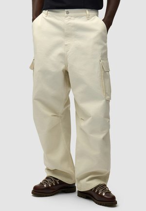 Uomo che indossa pantaloni cargo beige larghi con tasche laterali e scarponi da trekking marroni, con le mani nelle tasche, in piedi su uno sfondo semplice.