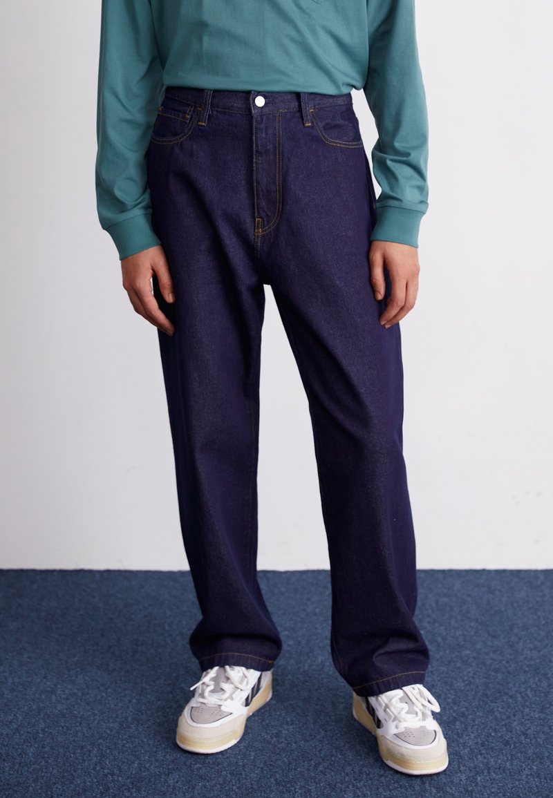 Mörk denimjeans med gul söm, lös passform, fem fickor, bärs med en långärmad teal skjorta och vita sneakers.