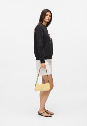 Donna con felpa nera e gonna bianca che tiene una piccola borsa gialla, indossa sandali infradito neri, in piedi su sfondo bianco.