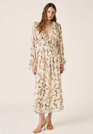 Mujer vestida con un vestido largo crema con estampado floral, escote en V con volantes, mangas largas con lazos y sandalias de tacón beige, de pie sobre un fondo liso.