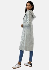 Lunga cardigan grigia a maglia con cappuccio, caratterizzata da un motivo a trecce e polsini e orlo a coste. Indossata con jeans blu e sneakers bianche.