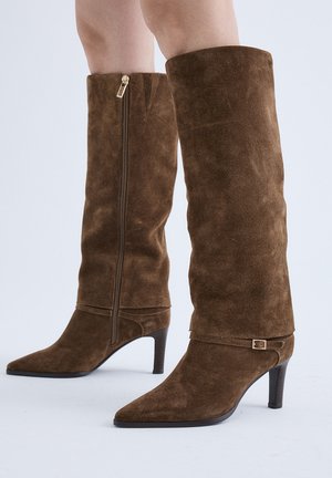 Botas marrones de ante hasta la rodilla con punta afilada, tacones de altura media, cremallera lateral y pequeño detalle decorativo con hebilla.