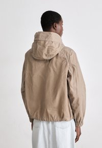 Beige színű, könnyű dzseki kapucnival, rugalmas mandzsettákkal és laza szabással. Simább textúrájú, egyszerű, minimalista dizájnnal rendelkezik.