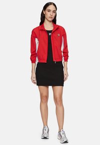 Veste coupe-vent rouge avec des accents blancs et bleus, robe noire ajustée et chaussures de sport grises. La veste a une fermeture éclair et des poignets élastiques.