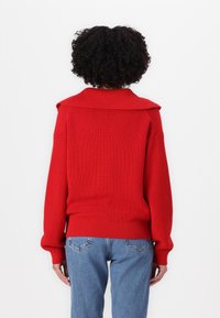 Rød strikket genser med bred krage, ribbestruktur og løs passform, kombinert med blåjeans. Bakken visningen fremhever genserens struktur.