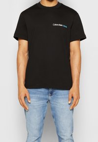 Černé bavlněné tričko s krátkým rukávem, kulatým výstřihem a malým modro-bílým logem "Calvin Klein Jeans" na levé části hrudi.